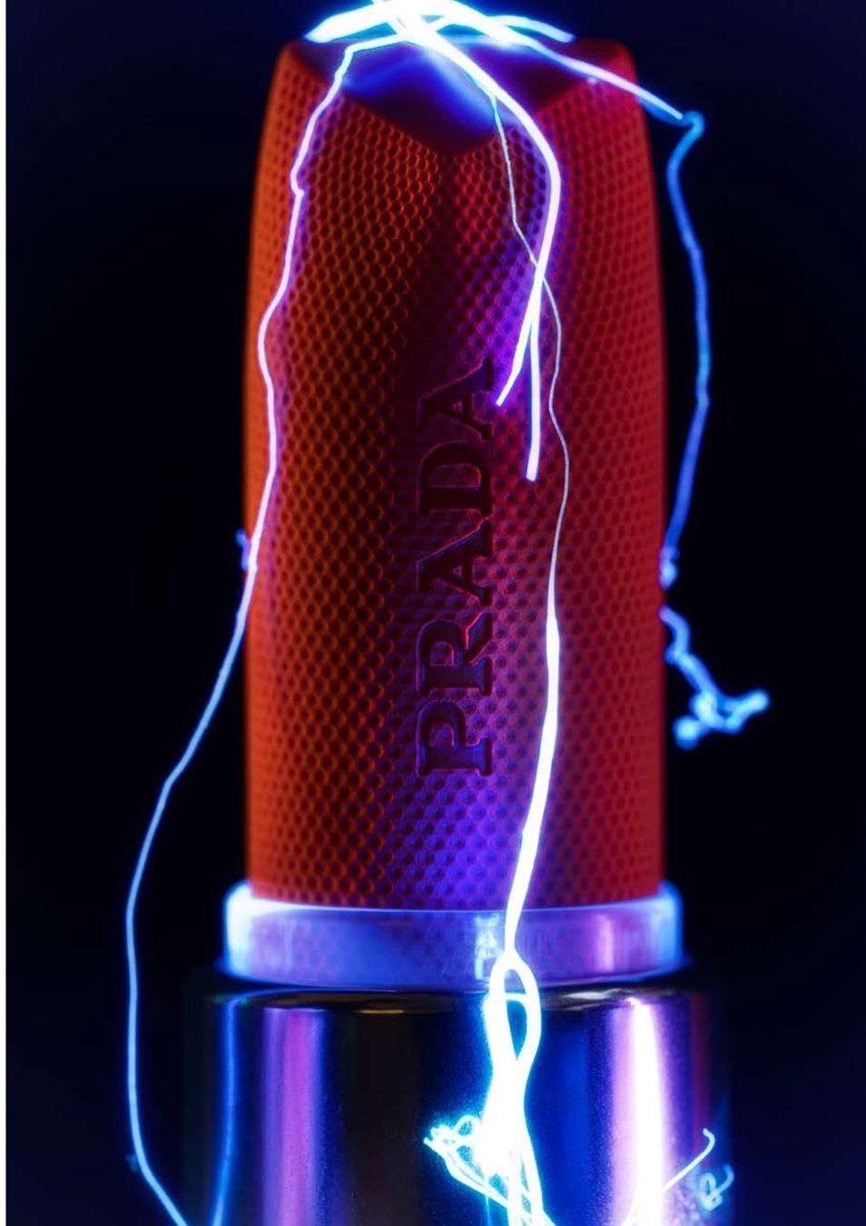 Still life featuring a red textured perfume bottle with blue neon lights and the Prada logo against a dark backdrop.