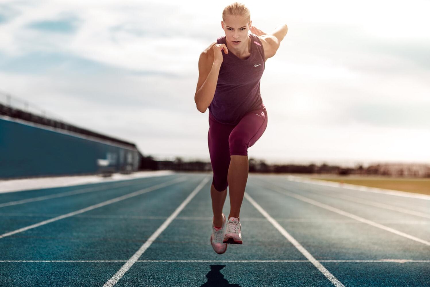 An athletic woman in running attire sprints forward on a blue running track beneath a partly cloudy sky, showcasing fitness and determination.