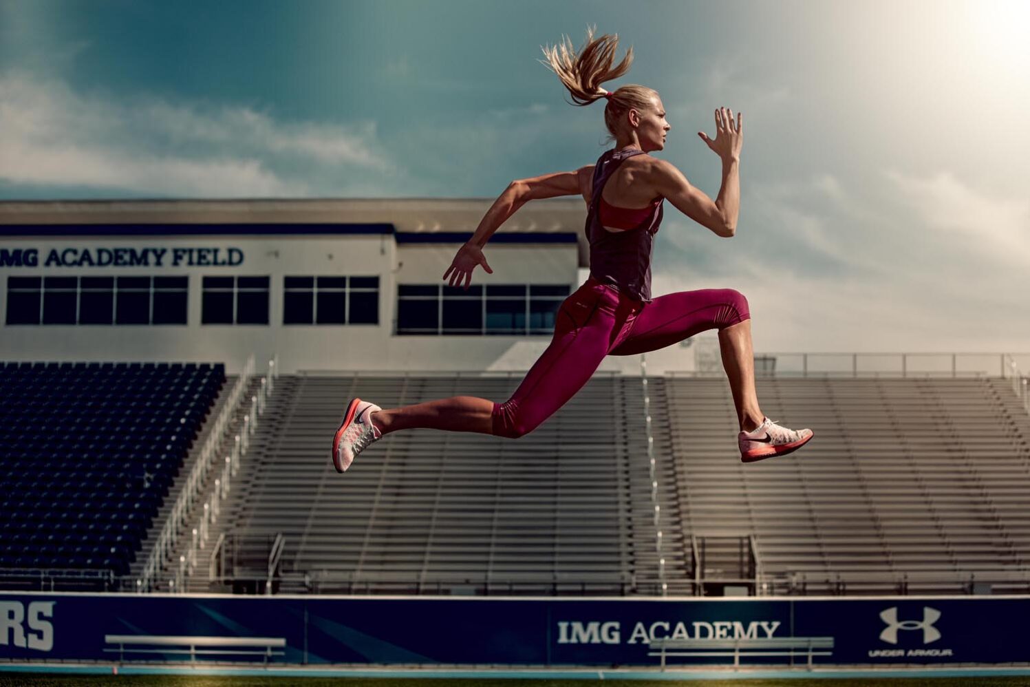 Athletic woman in mid-air running on a track with stadium seating and blue sky in the background, showcasing fitness and determination.