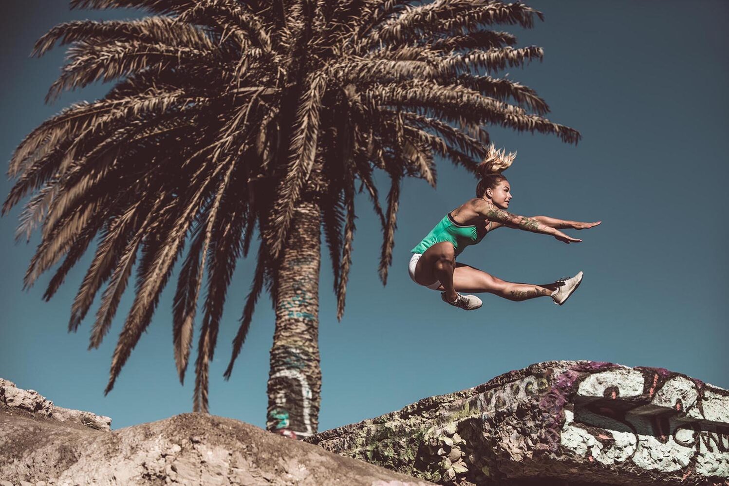Athletic woman in sportswear jumps in the air near a large palm tree against a vibrant blue sky, showcasing fitness and outdoor activity.