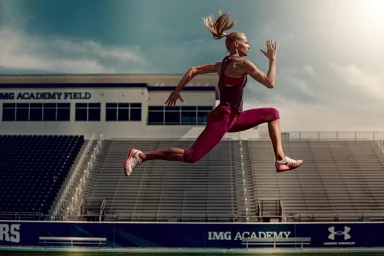 Blonde female athlete in burgundy sportswear leaping mid-air across an outdoor sports stadium track.