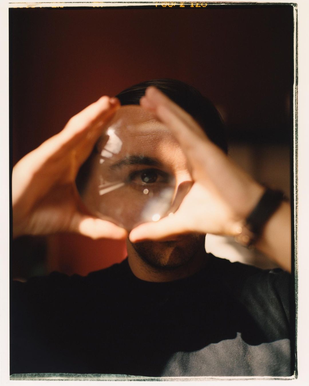 Man framing face with hands around a clear glass ball, wearing a black t-shirt and a watch, indoors against a dark red wall.