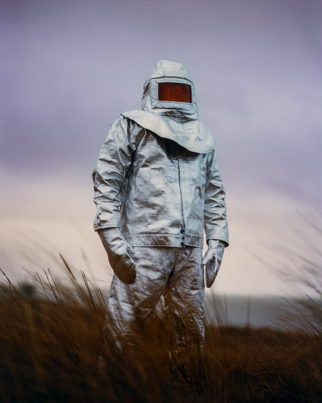 Person in a silver proximity suit standing in a field of tall grass, against a sky of purple and gray tones, facing forward.