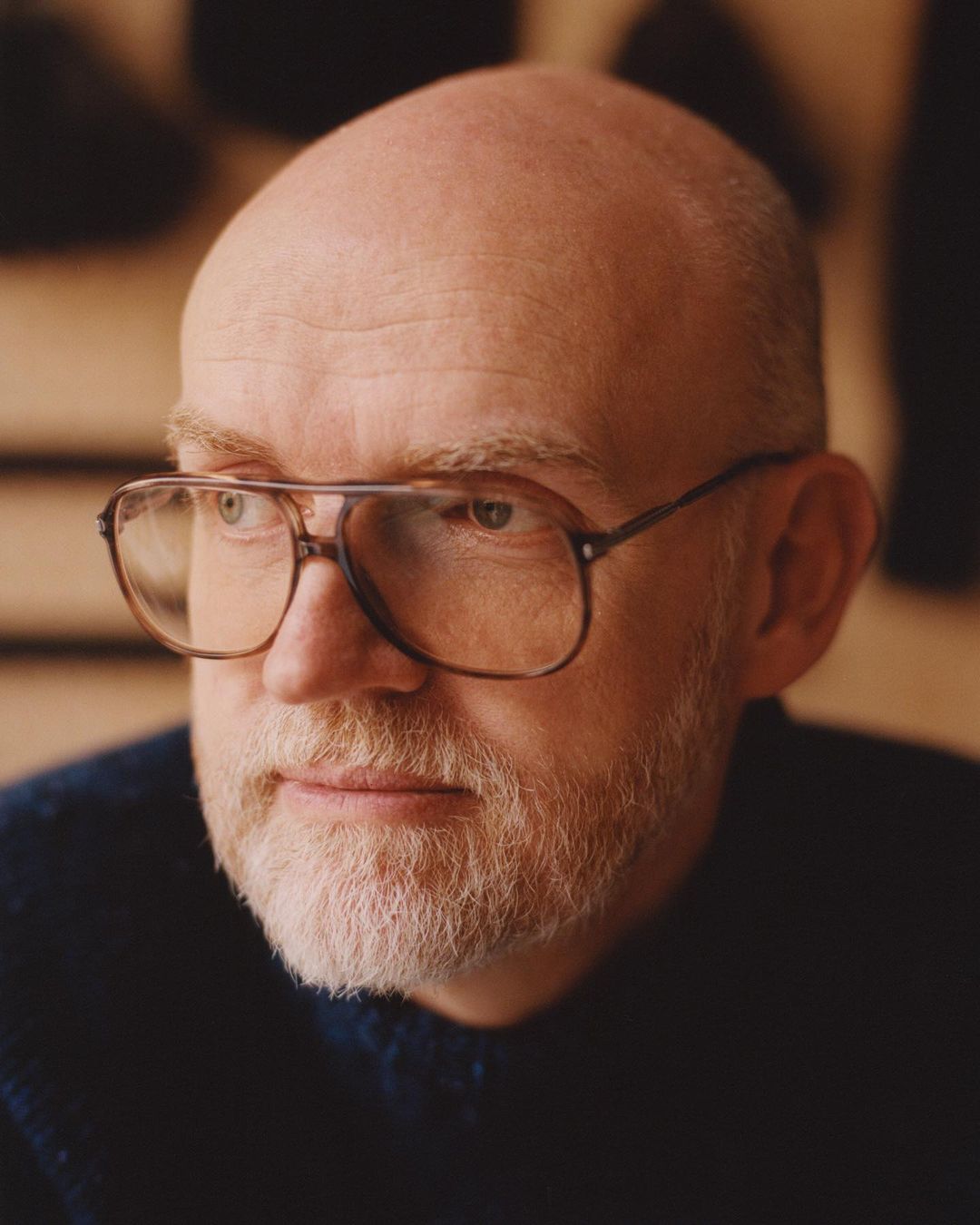 Close-up portrait of a balding, mature man with a beard wearing glasses, against a blurred background with soft, diffused lighting.