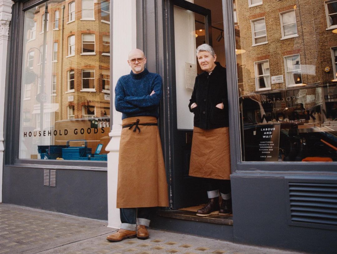 Two elderly business owners stand in the doorway of their household goods store on a city street; natural light, exterior shot.