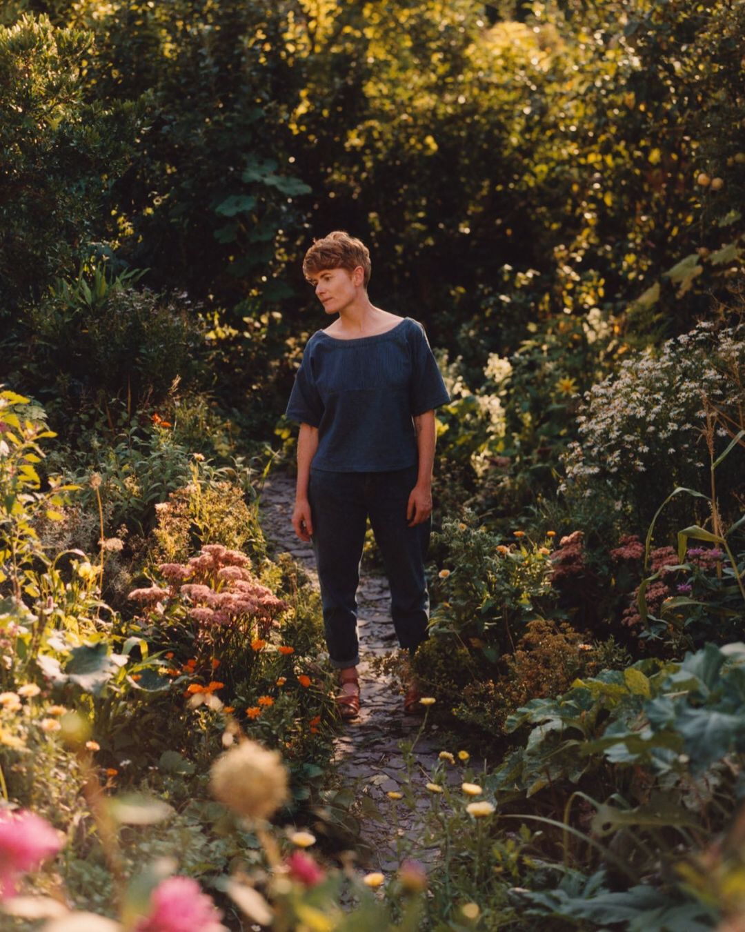 Woman in casual blue outfit stands on a stone path in lush garden with abundant flowers, surrounded by greenery and dappled sunlight.