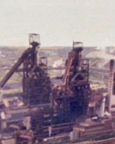 Industrial scene featuring large, weathered steel structures, possibly blast furnaces, against a muted, slightly blurred urban backdrop.