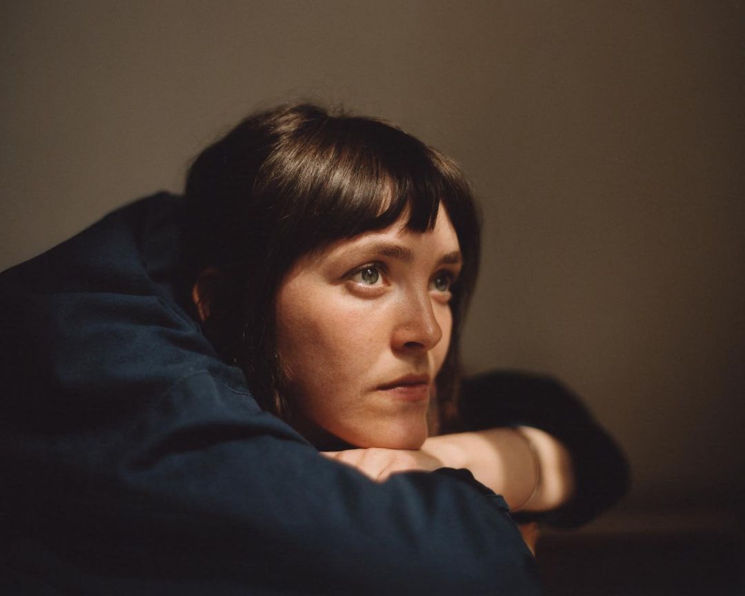 Portrait of an adult female with dark hair resting her head on her hands looking up in an indoor setting with soft, warm lighting.