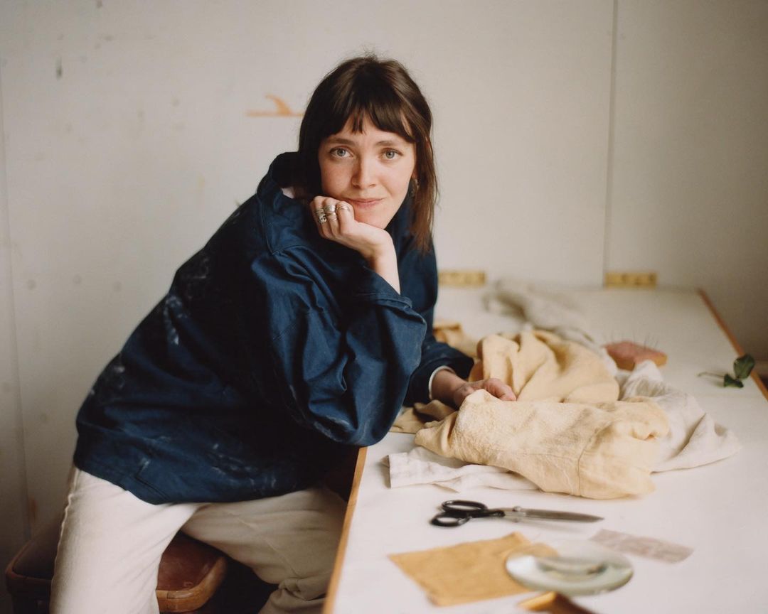 Woman sits at a table in a relaxed pose, wearing a blue jacket, surrounded by textiles, scissors, and a dish, in an indoor setting.