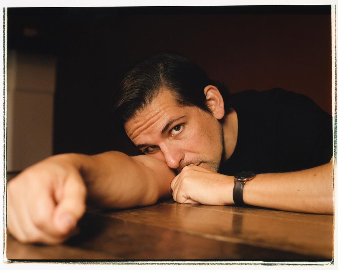 Portrait of a man resting his face on his arm while lying on a brown table, wearing a black shirt and a wristwatch indoors.