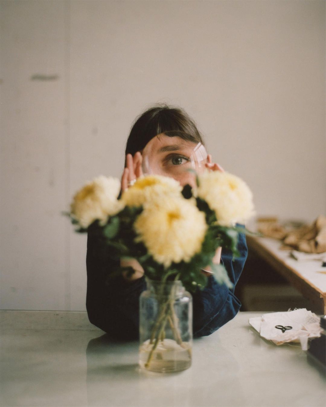 Adult female partially visible behind a clear glass vase filled with yellow chrysanthemum flowers, sitting atop a white table indoors.