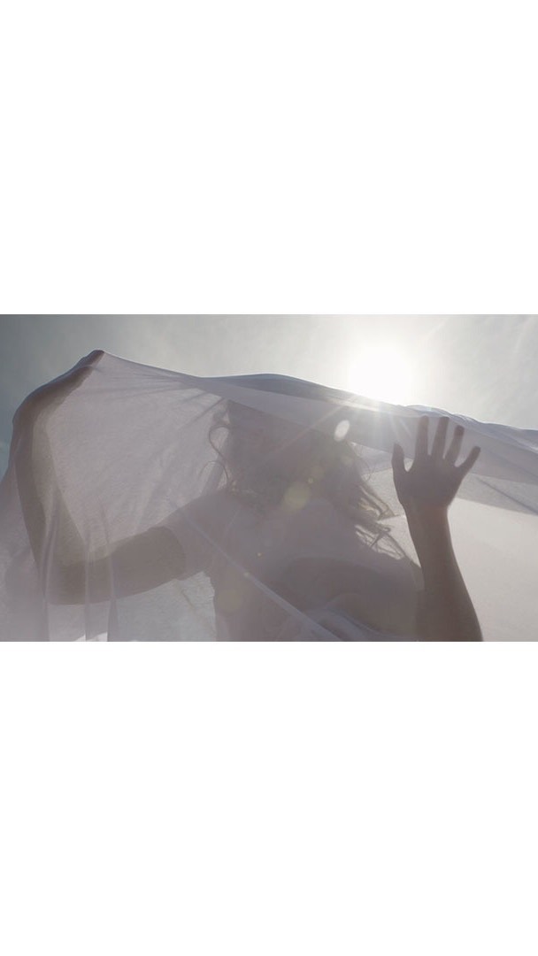 Woman stands outdoors backlit, covered by a translucent white fabric that she holds up with both hands against a bright sky.