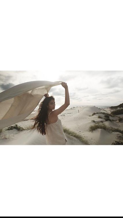 Young woman in a light dress stands on a sandy beach under an overcast sky, holding a translucent cloth overhead. Shot from behind.