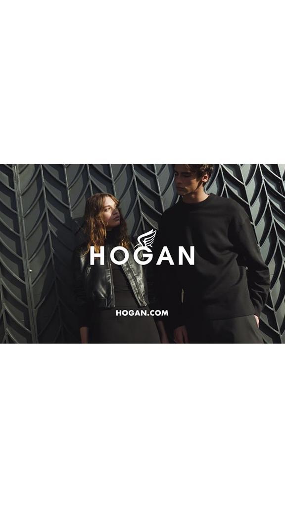 A stylish couple posing against a patterned wall, the HOGAN logo and website address prominently displayed, in a fashion-forward editorial shot.