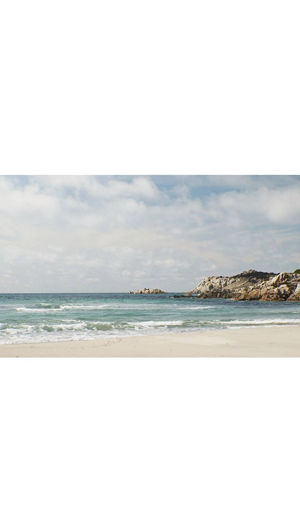 Beach scene featuring a white sandy shore meeting the blue ocean water under a partly cloudy sky, with rocky cliffs in the background.