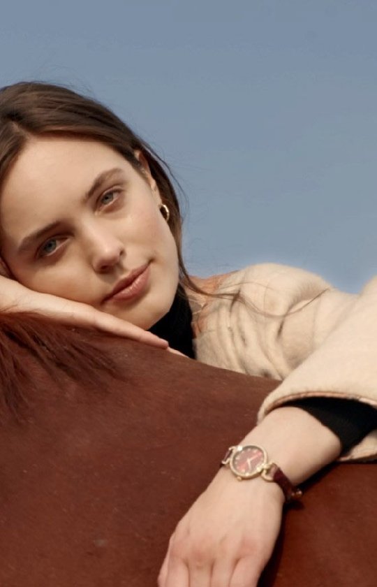 Portrait of a woman with brown hair leaning on a brown horse outside on a sunny day, wearing a beige jacket and a wristwatch.