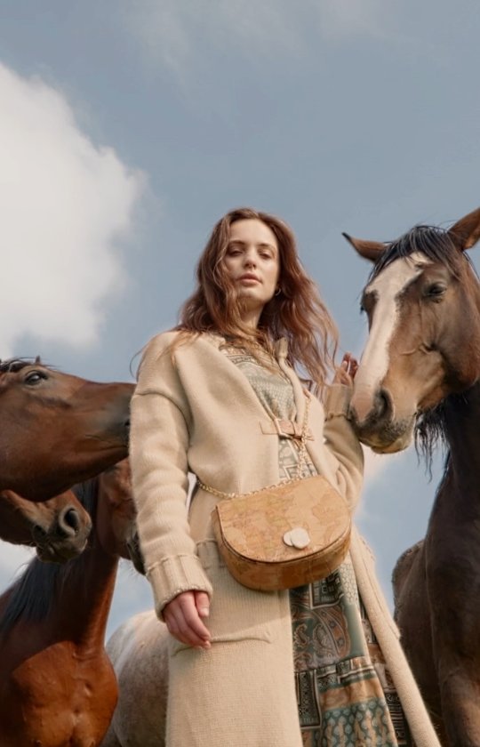 A woman in a coat with a purse stands in a field surrounded by horses under a partly cloudy sky in a fashion editorial.