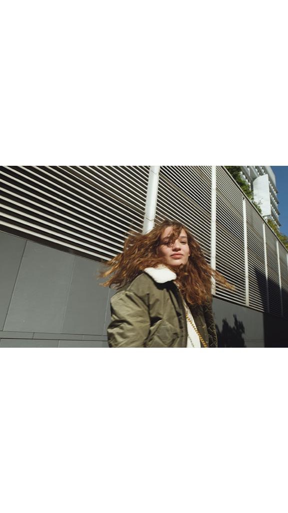 Woman with wavy red-brown hair in an olive green jacket poses in front of an urban building with horizontal metal slats.