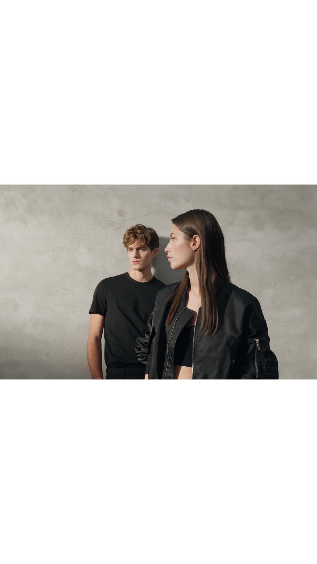 A dark-haired woman in a jacket and a light-haired man in a black t-shirt pose in front of a textured gray wall indoors.