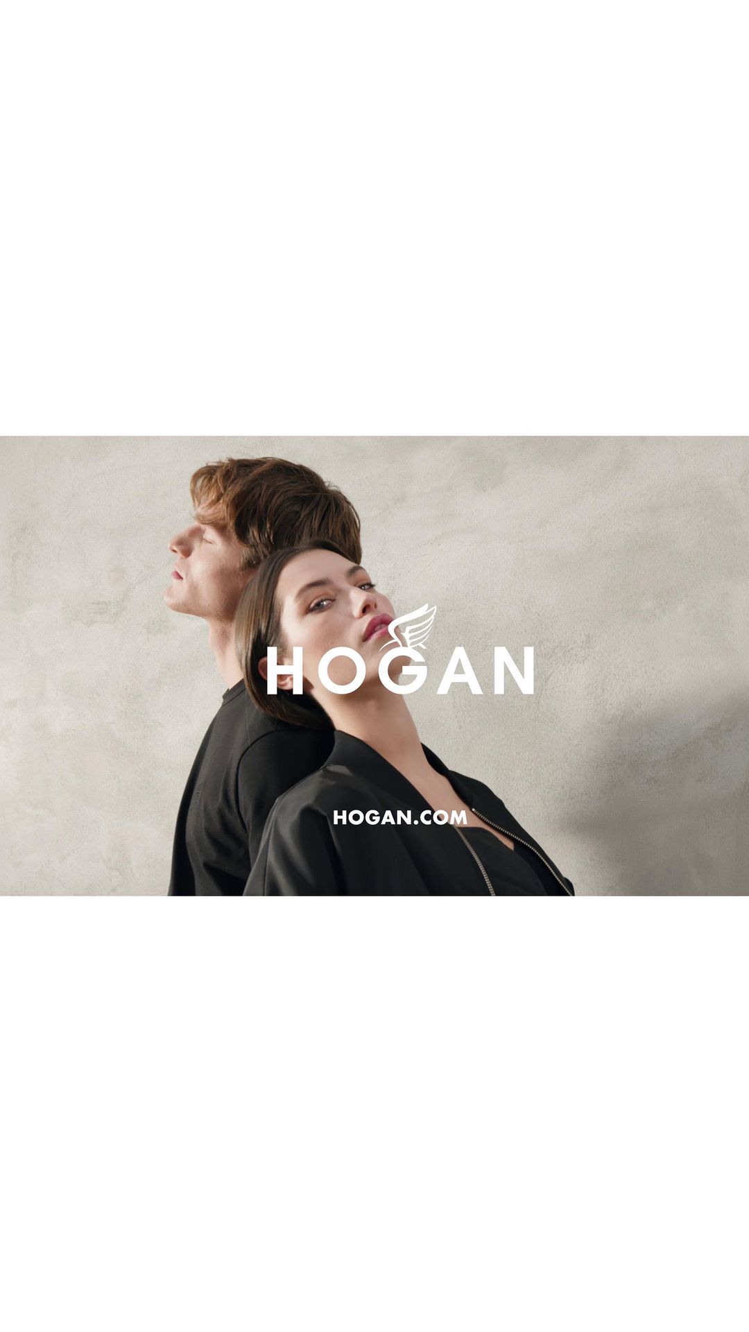 Two models strike a pose in a promotional shot for HOGAN, set against a textured gray backdrop. The brand's logo is prominently displayed.