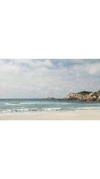 A wide sandy beach meeting gentle turquoise ocean waves under a partly cloudy sky with rocky outcrops in the distance.
