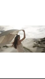 Woman holding a white fabric in the wind amidst sand dunes under an overcast sky.
