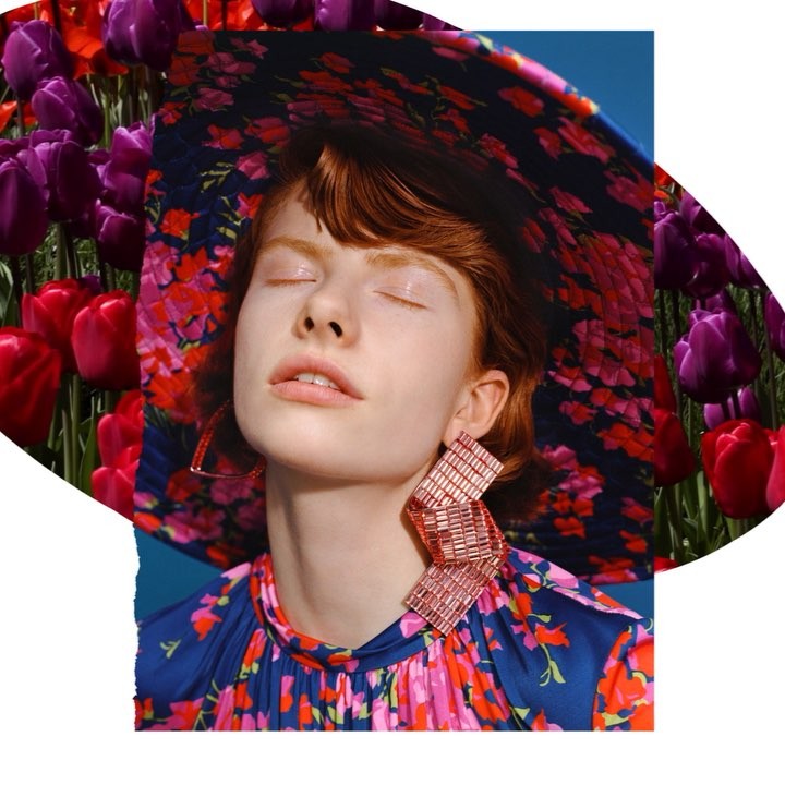 Adult woman with red hair and closed eyes, wearing a floral hat and large earrings, surrounded by red and purple tulips.