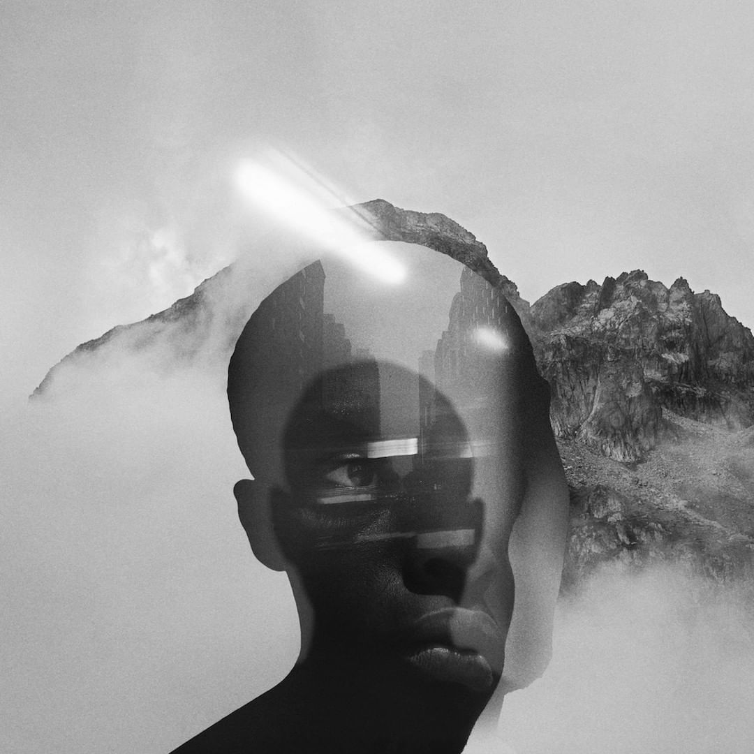 Monochrome portrait with double exposure of a person's face, a cityscape, mountains, and fog creates an abstract, surreal atmosphere.