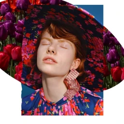 Close-up portrait of a red-haired woman wearing a floral wide-brimmed hat and geometric earrings against a tulip garden background.