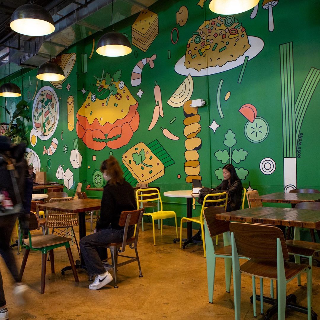 Indoor cafe scene with two women at tables and a bright mural featuring whimsical illustrations of food dishes in vibrant colors.
