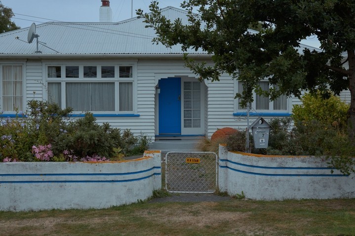 Exterior view of a white single-family home with a blue door, a small front yard with flowers, and a white wall with blue trim.