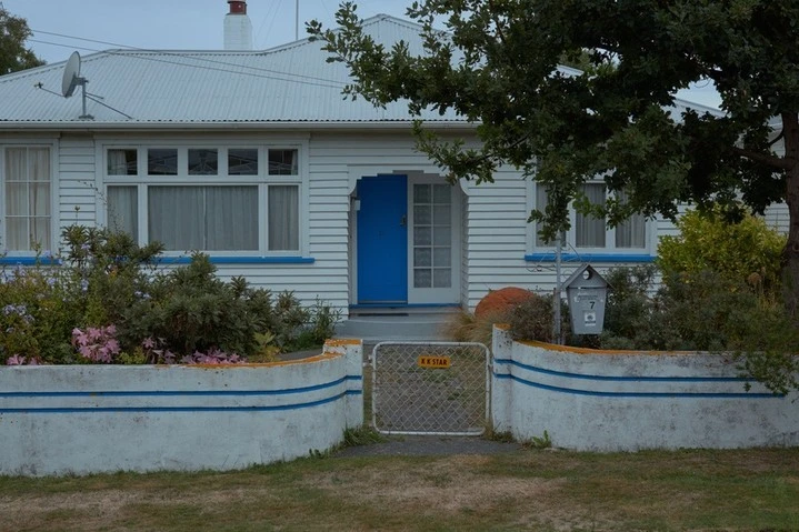 Exterior view of a white single-family home with a blue door, a small front yard with flowers, and a white wall with blue trim.