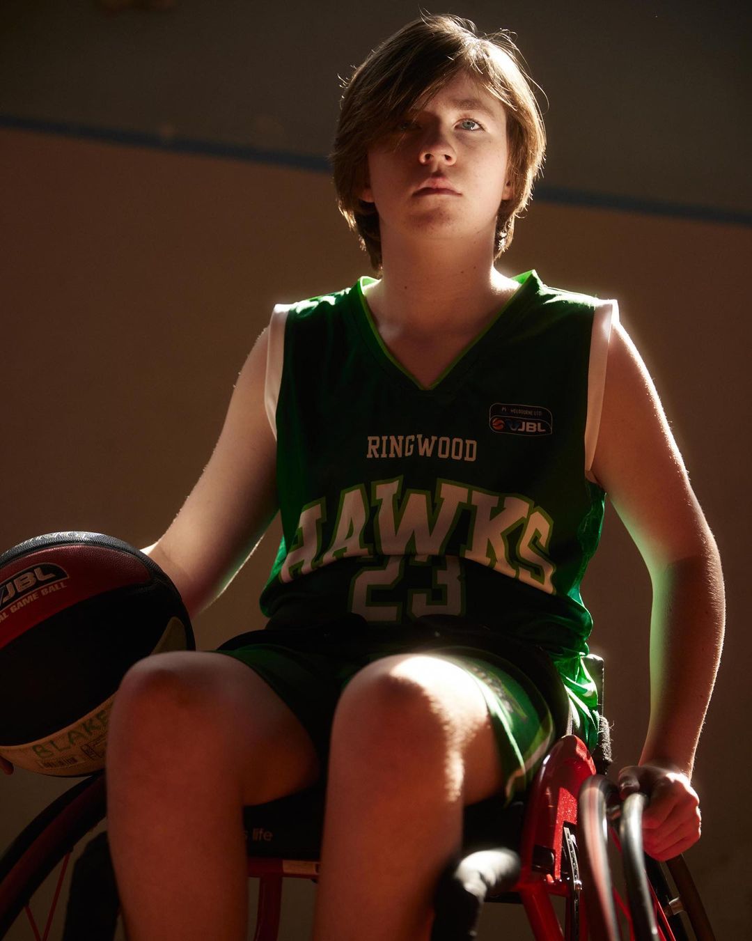 Teen boy in wheelchair wearing basketball jersey, holding a basketball, indoors, youth sports.