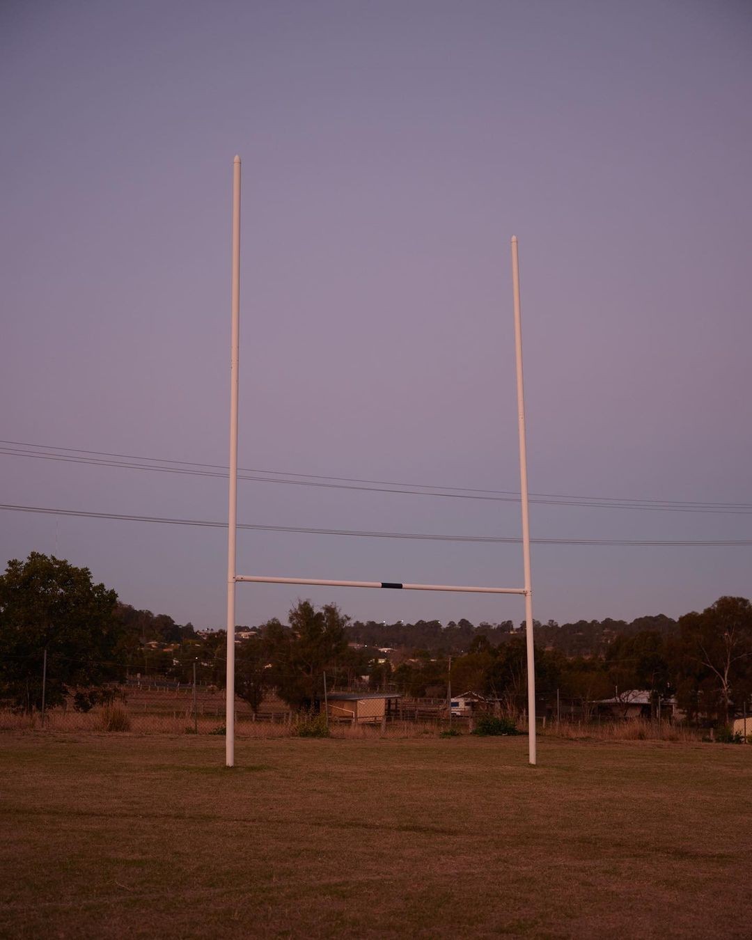 Rugby goal posts stand on a grassy field at dusk, with a tree-lined horizon and soft lavender sky, in a rural setting.