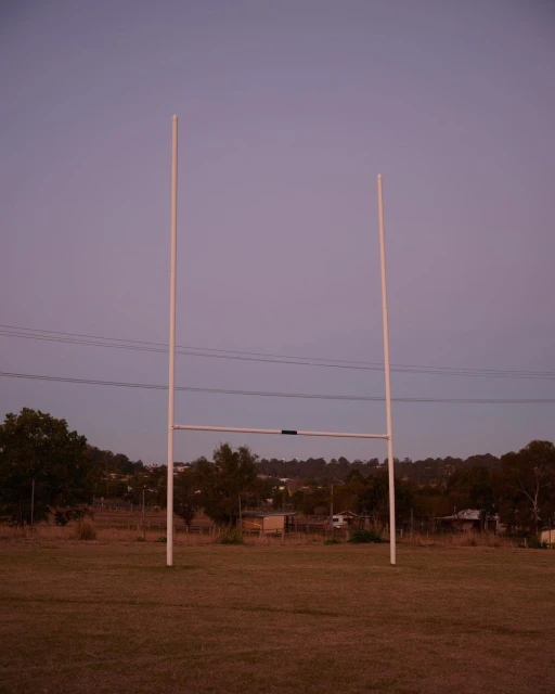 Rugby goal posts stand on a grassy field at dusk, with a tree-lined horizon and soft lavender sky, in a rural setting.