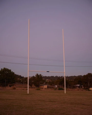 Rugby goal posts stand on a grassy field at dusk, with a tree-lined horizon and soft lavender sky, in a rural setting.