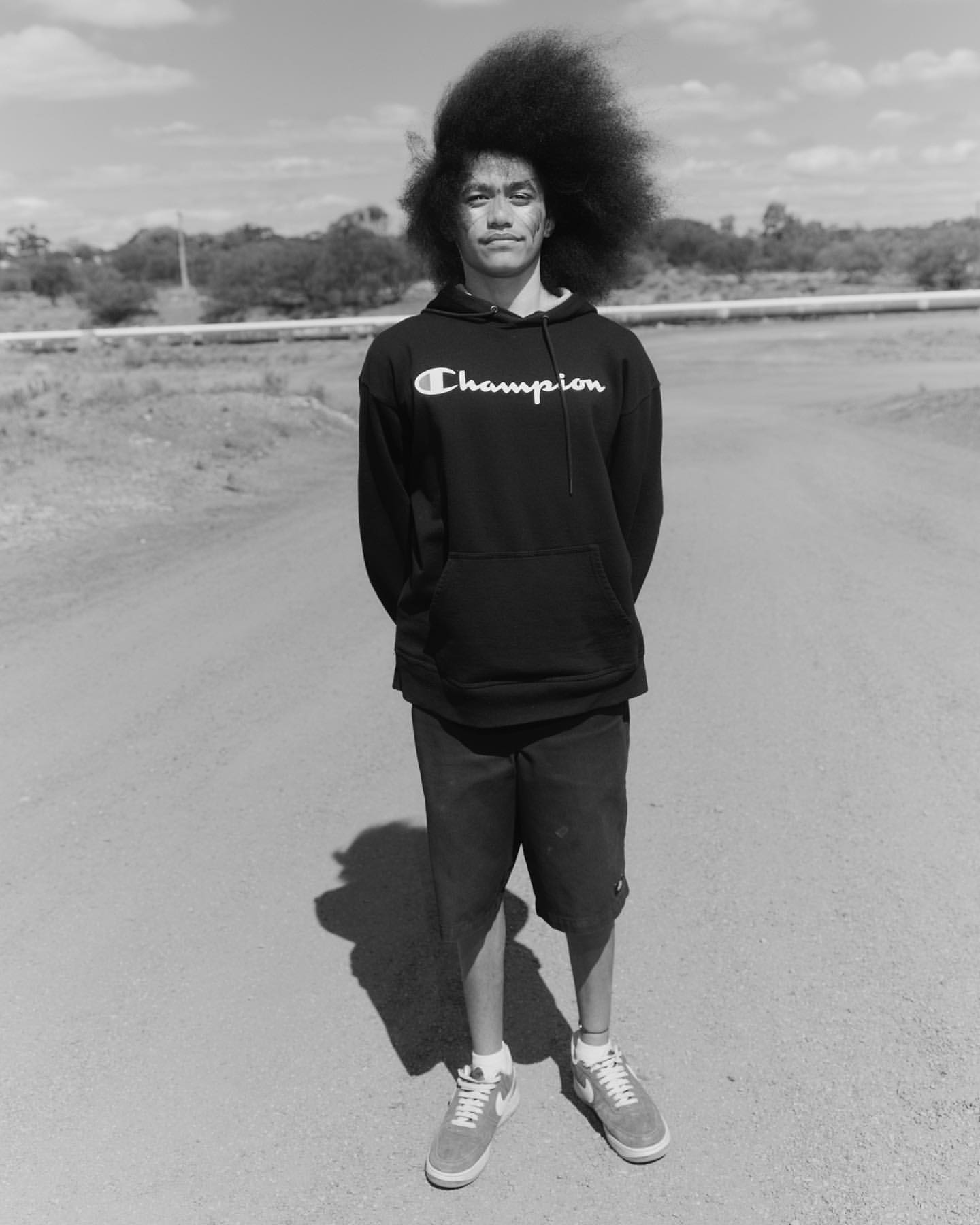 Monochromatic portrait of a young adult man with an afro hairstyle standing on a dirt road wearing a Champion hooded sweatshirt.