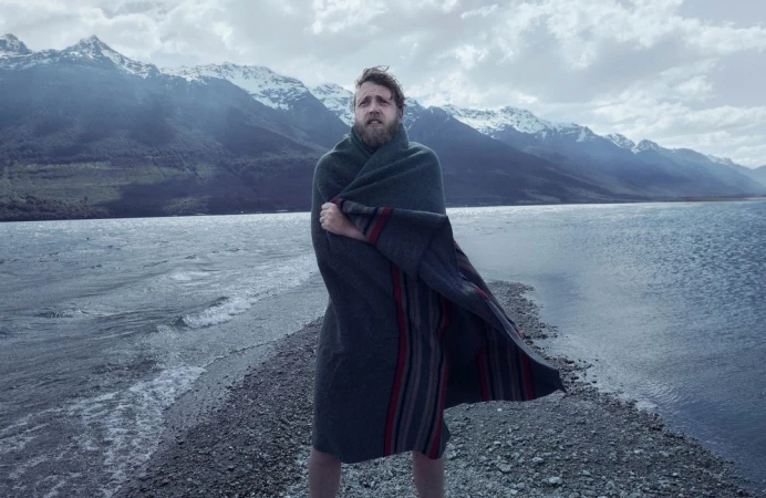 Bearded man wrapped in a dark woolen blanket standing on a gravel spit by a lake with snowy mountains.