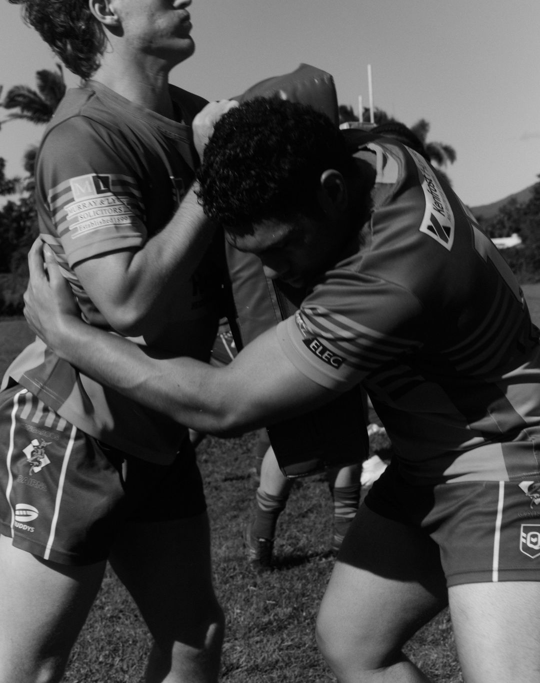 Two male rugby players grapple on a grassy field under bright sunlight, showcasing their strength and teamwork in a monochrome image.