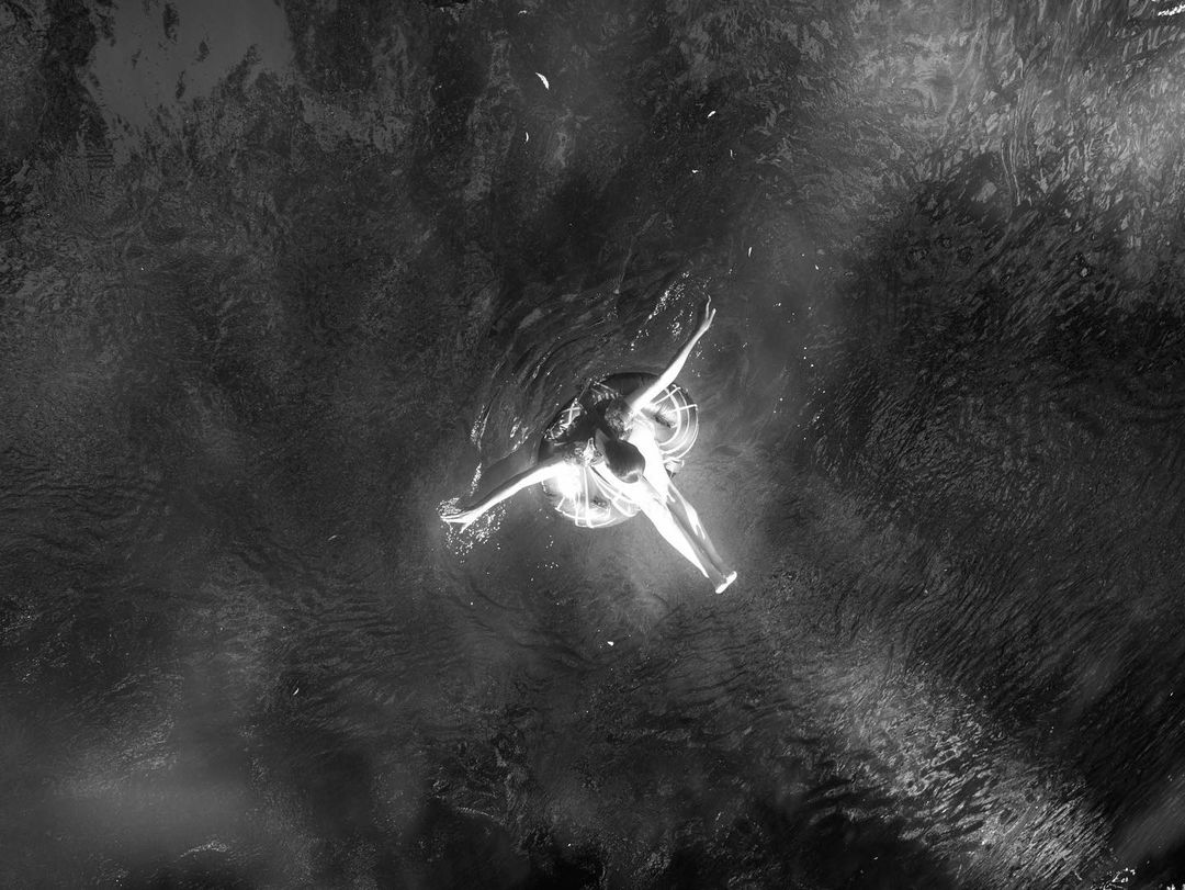 Monochrome image of an adult female floating in water on a ring float, captured from an overhead angle in a ripple-filled scene.