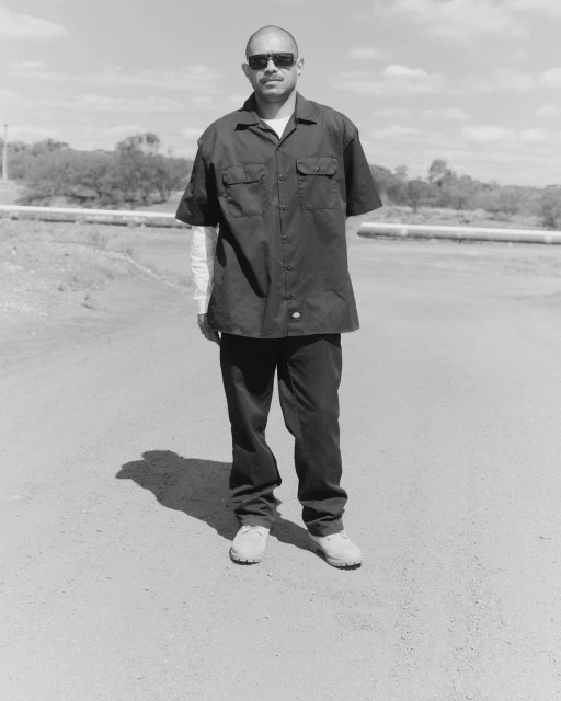 Monochrome portrait of a man in sunglasses and work clothes standing on a road, set against a sky and industrial backdrop.