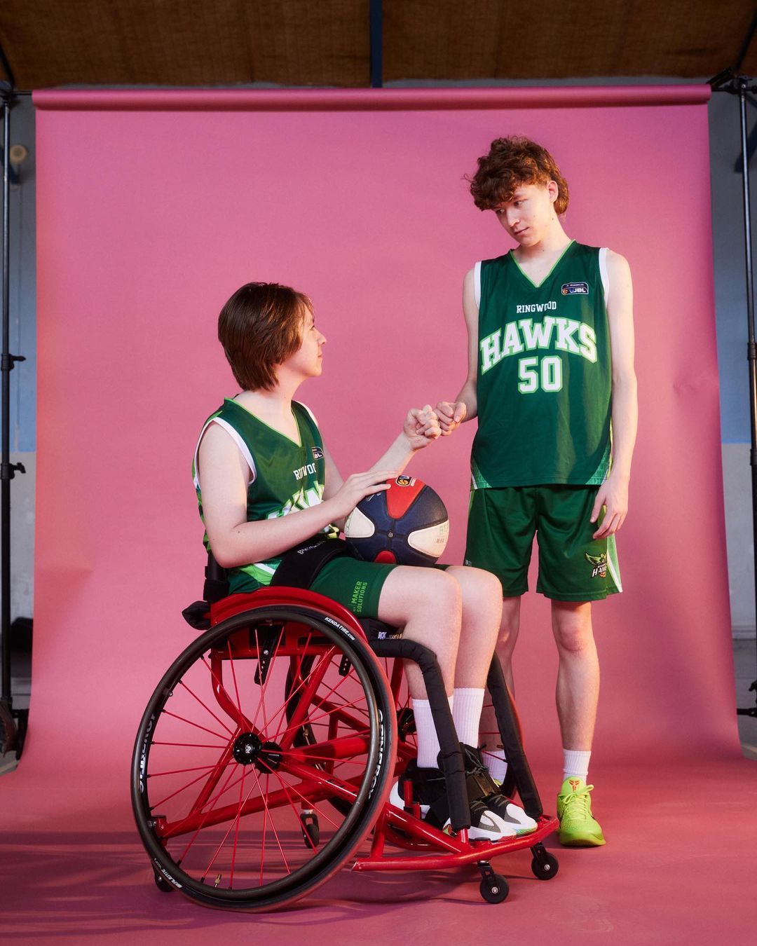 Two basketball players, one in a wheelchair, on a pink backdrop. Athlete in wheelchair holds a basketball, both in sports uniforms.