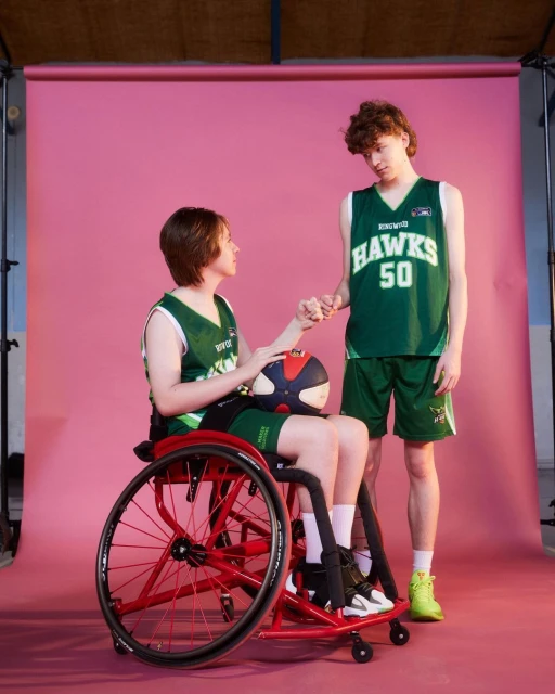 Two basketball players, one in a wheelchair, on a pink backdrop. Athlete in wheelchair holds a basketball, both in sports uniforms.