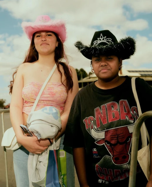 Two women pose outdoors wearing cowgirl hats and casual clothing, one in pink and the other in black, under a bright sky.