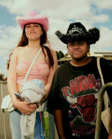 Two women pose outdoors wearing cowgirl hats and casual clothing, one in pink and the other in black, under a bright sky.