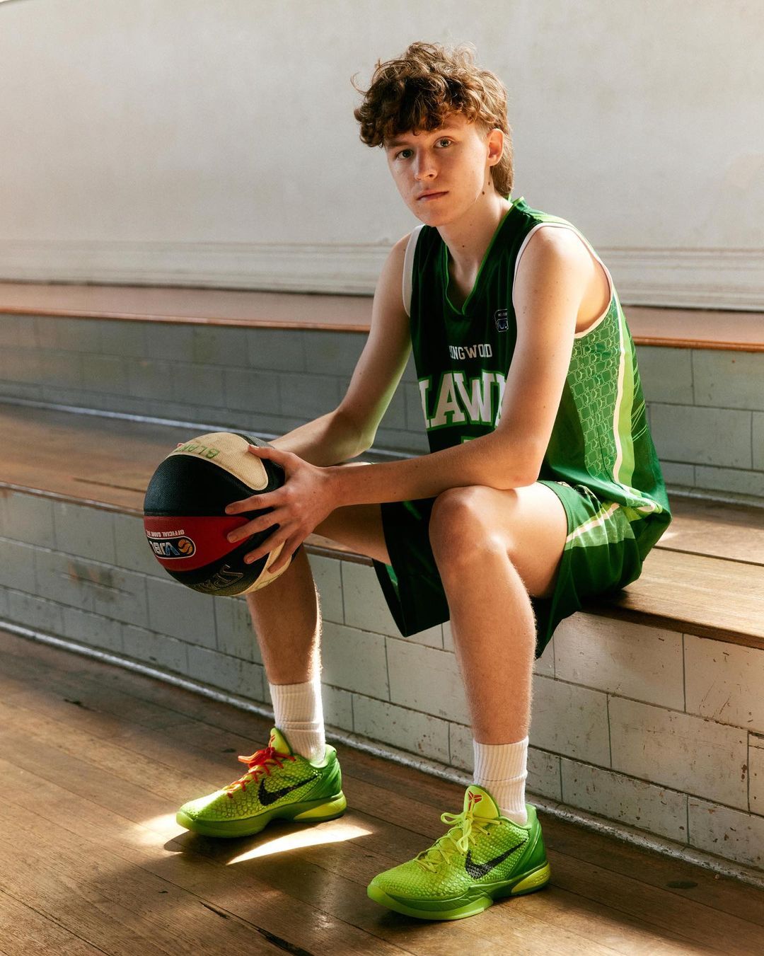 A young adult male in a green basketball jersey sits with a basketball, wearing bright green sneakers on a hardwood surface indoors.