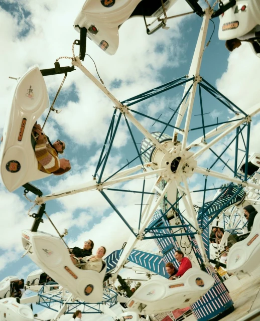 People ride a white carnival ride with blue accents against a cloudy sky, capturing a low-angle view emphasizing the ride's height and motion.