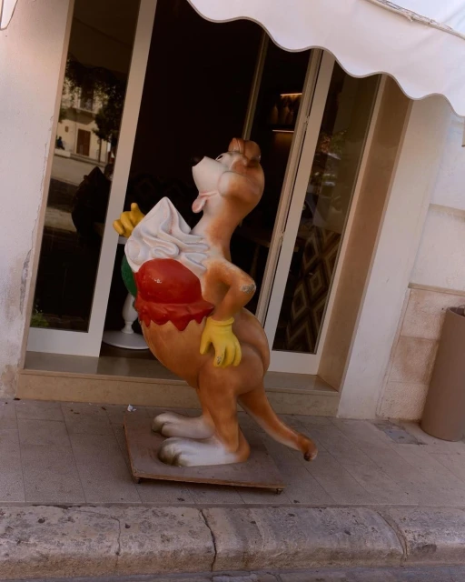 A fiberglass kangaroo statue holds an ice cream cone outside a building, viewed at eye level on a sunny day.