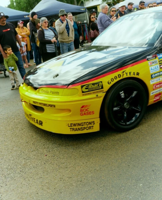 Yellow race car with black accents parked near a crowd of spectators outdoors under an overcast sky during the daytime.