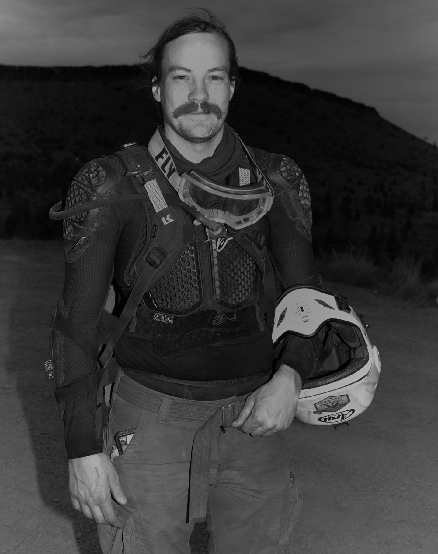 Portrait of adult male motorcyclist with helmet in hand, wearing protective gear, outdoors in a rural mountain landscape in black and white.